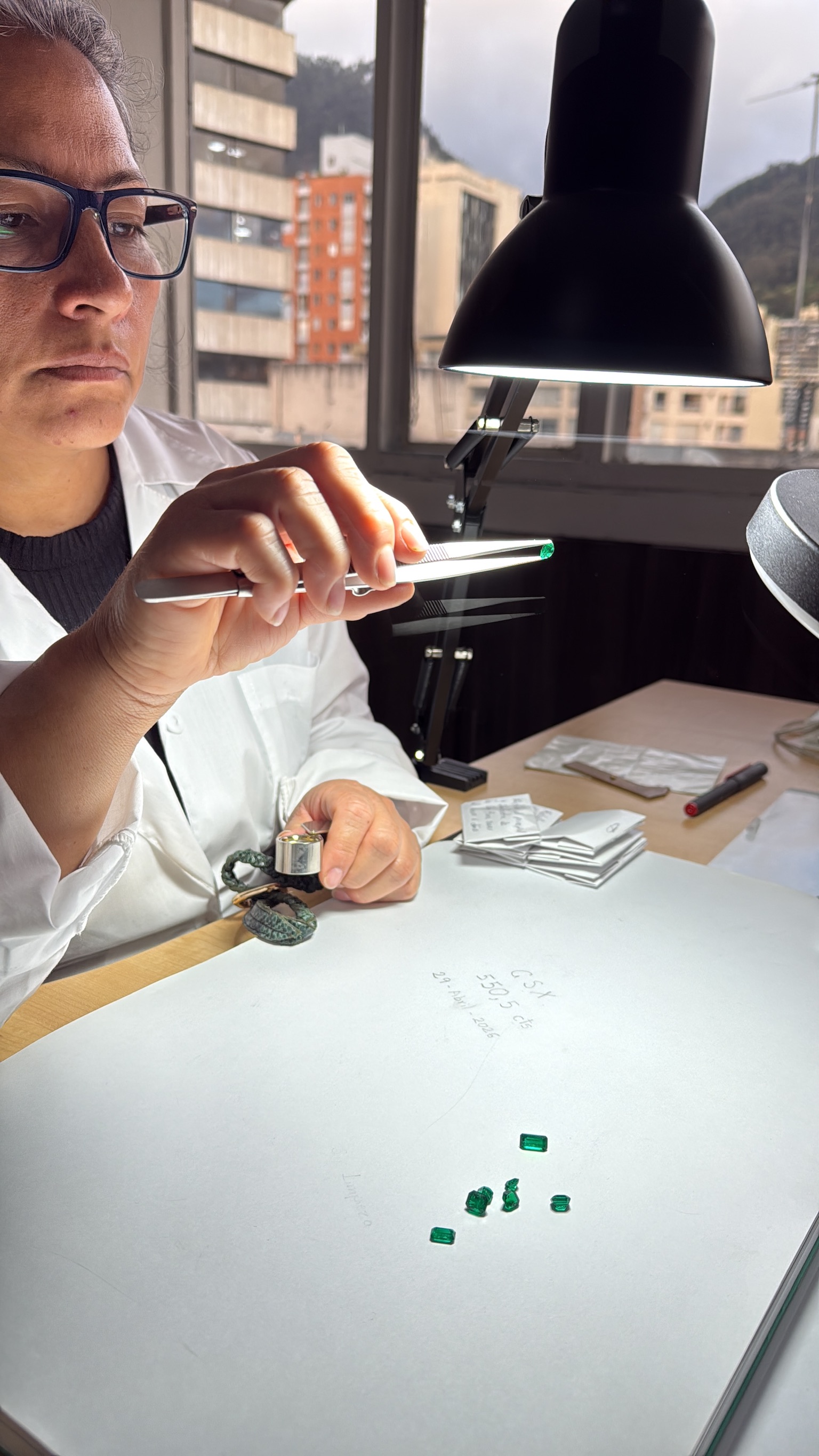 A GemstoneX gemmologist grading loose Colombian emeralds at the loupe in the Bogotá office, the city visible through the window